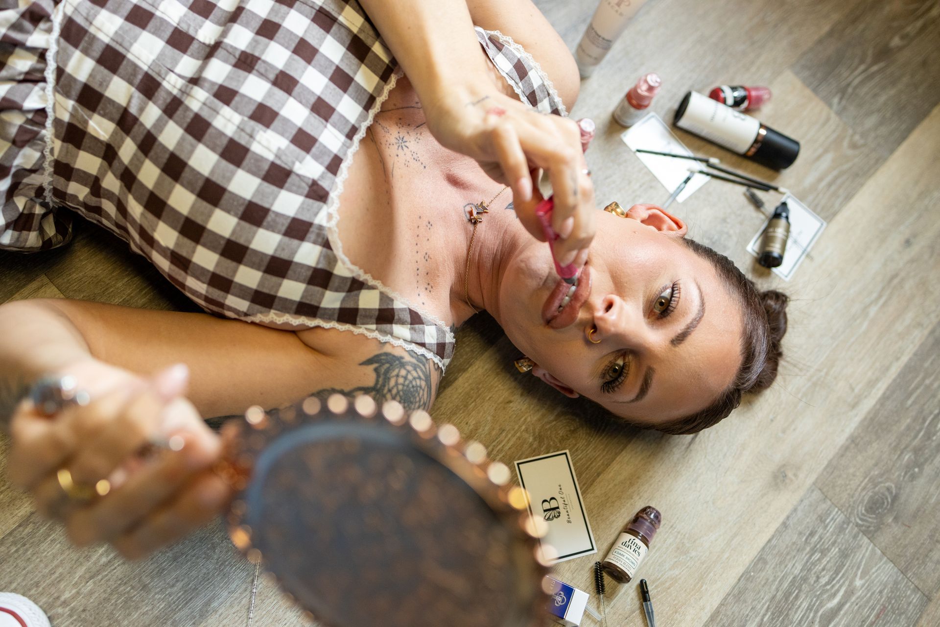 Woman applying lipstick while holding a mirror on a wooden floor, with makeup tools nearby