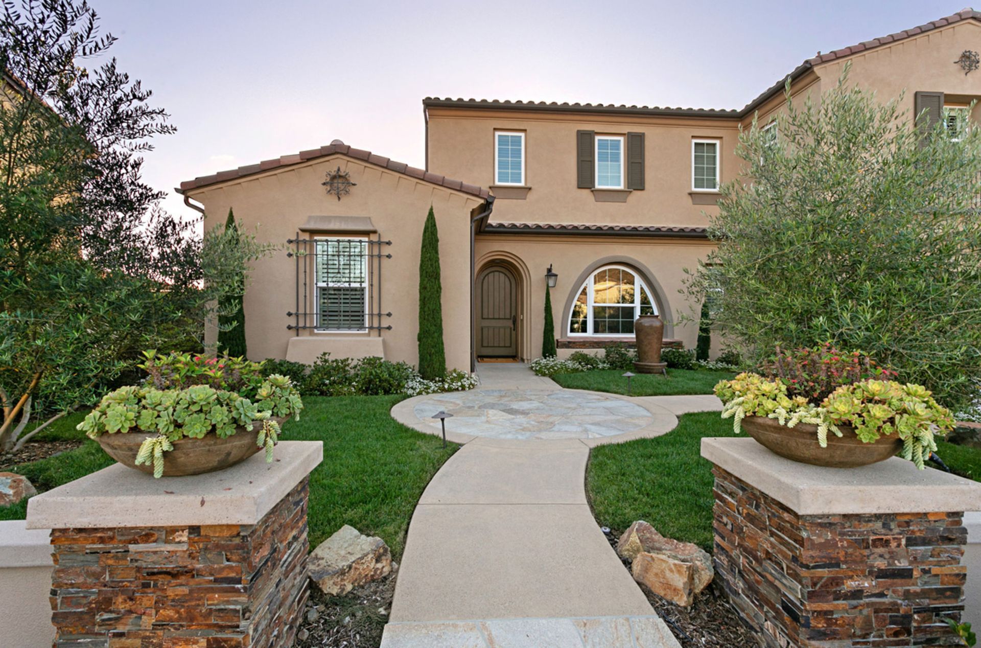 Two-story beige house with stone pathway and planters. Lush green lawn and mature trees in front.