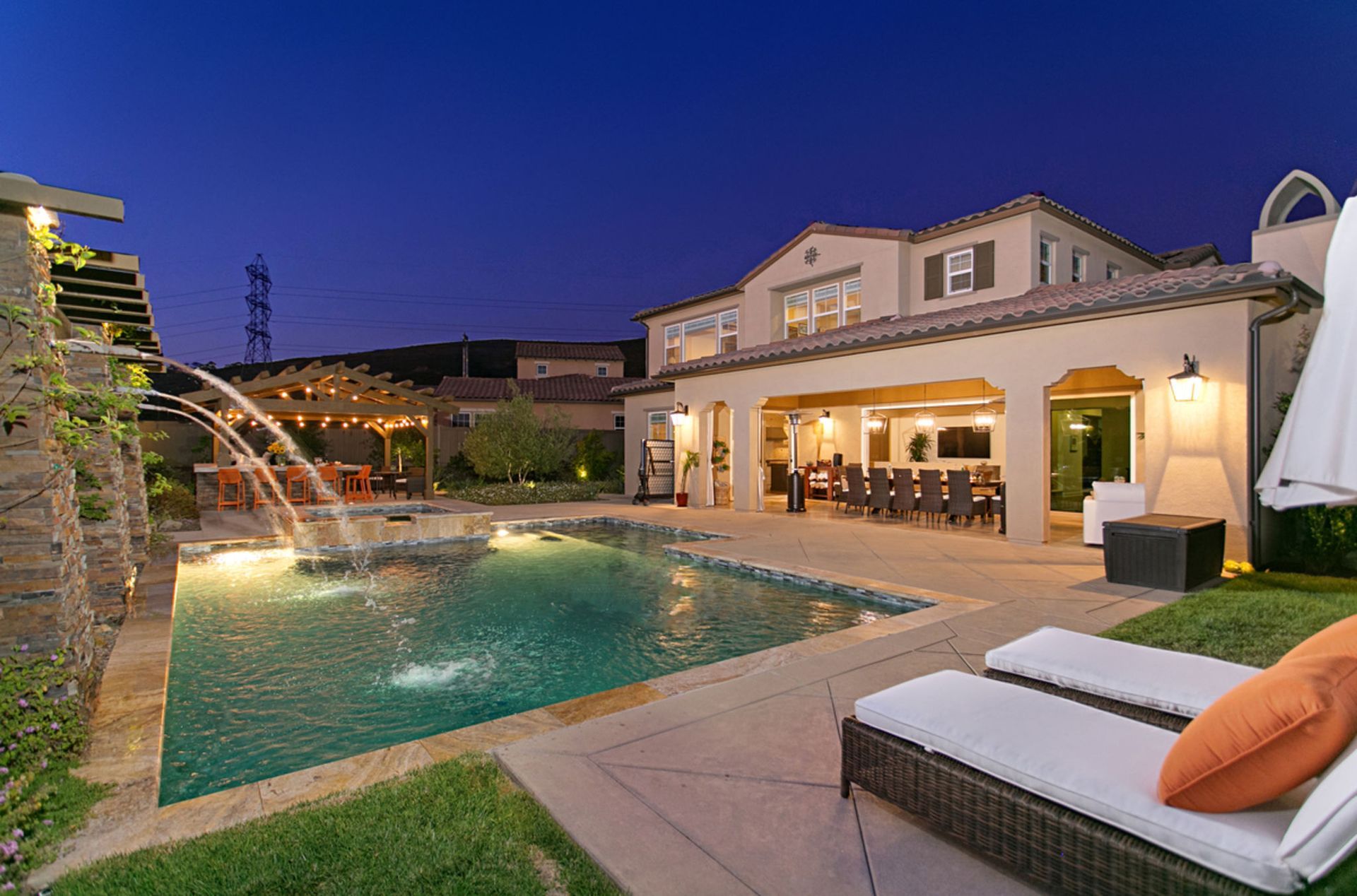 Backyard pool at dusk with a two-story house, fountain, and seating. The setting is lit with artificial lights and a dark blue sky.