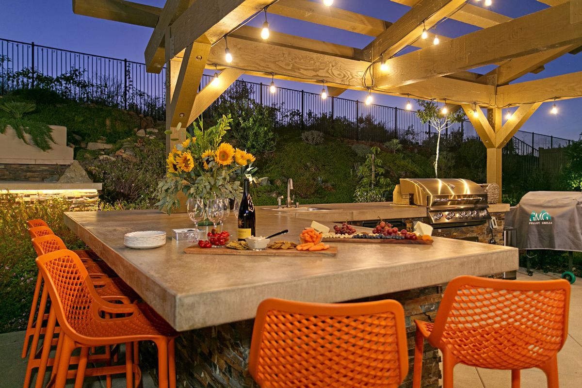 Outdoor kitchen with orange bar stools, food and drinks on the counter, and overhead string lights, at dusk.