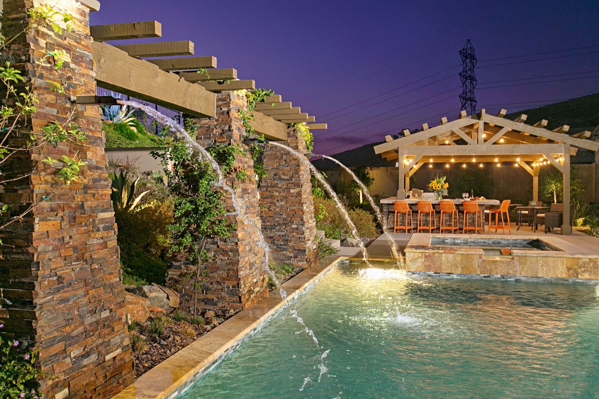 A luxurious backyard pool with a stone waterfall feature. A gazebo with a dining table and string lights sits nearby, under a purple twilight sky.