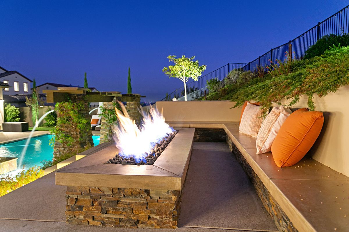 Outdoor patio with a built-in fire pit, seating, and colorful pillows. A pool and hillside are in the background at dusk.
