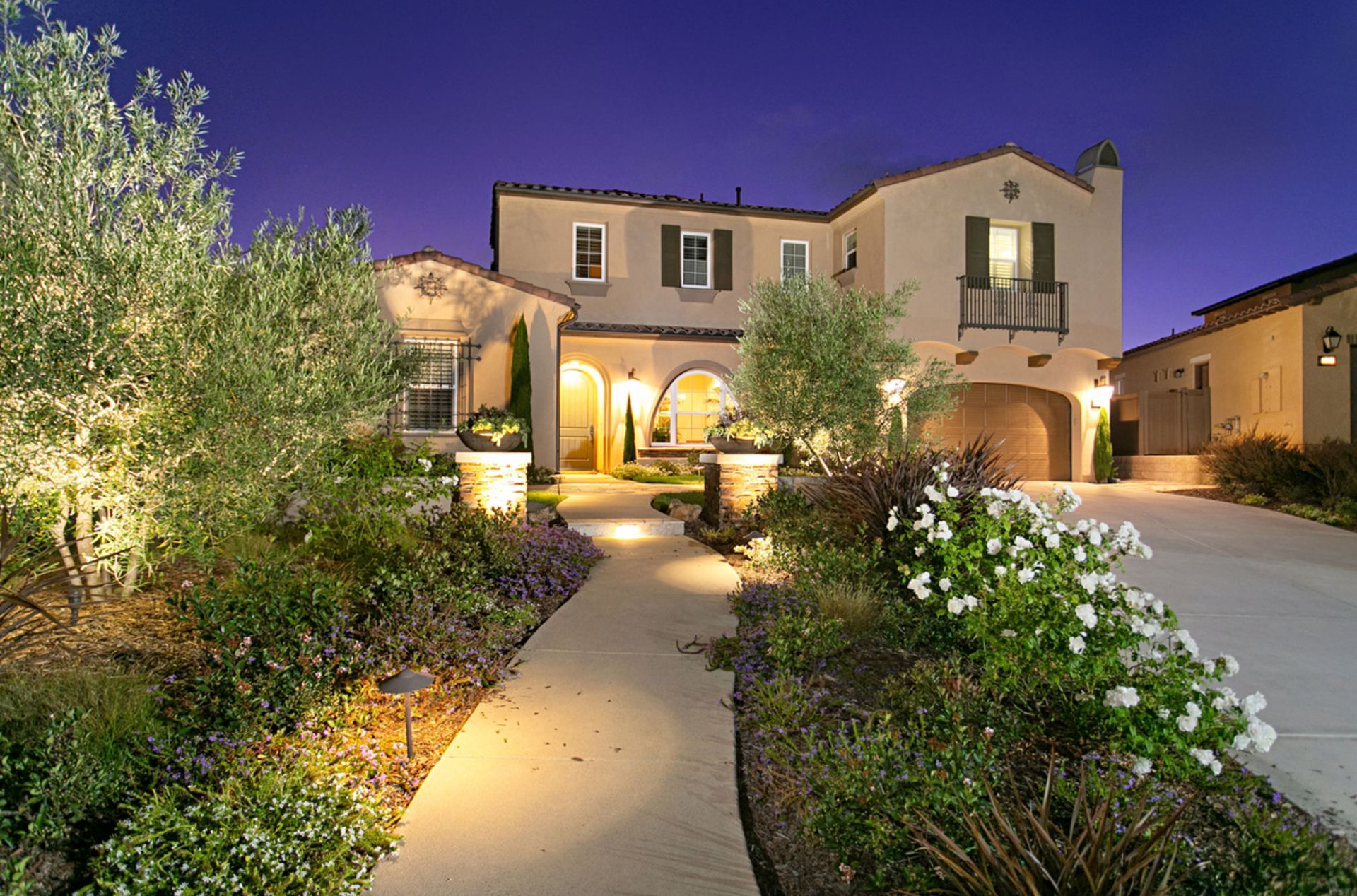 Two-story beige house with a lit pathway leading to the arched entrance. Landscaping and a driveway are visible in the twilight.