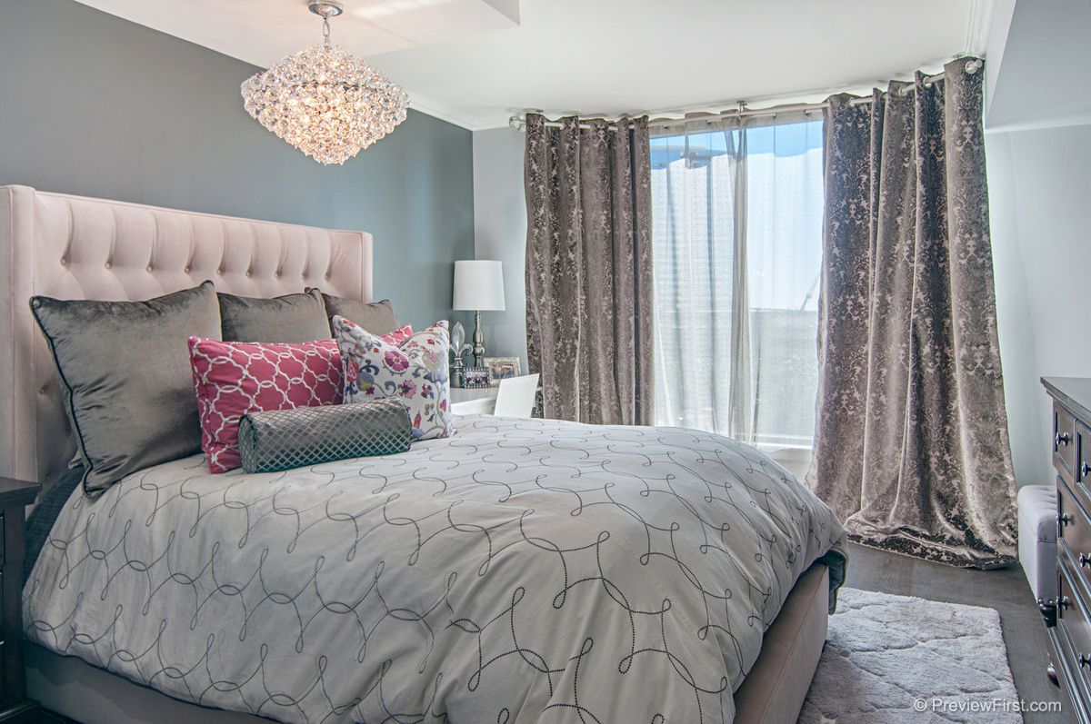 A luxurious bedroom with a large bed, pink headboard, and decorative pillows. The room features gray walls, elegant curtains, and a crystal chandelier.