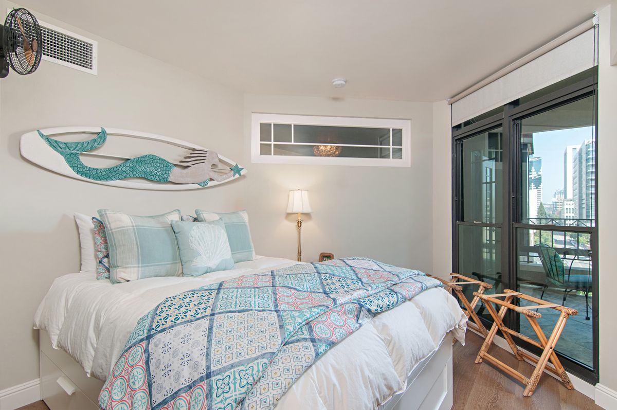 Bedroom with white walls, a double bed with blue and white bedding, and a surfboard with a mermaid design above it. Balcony doors are to the right.