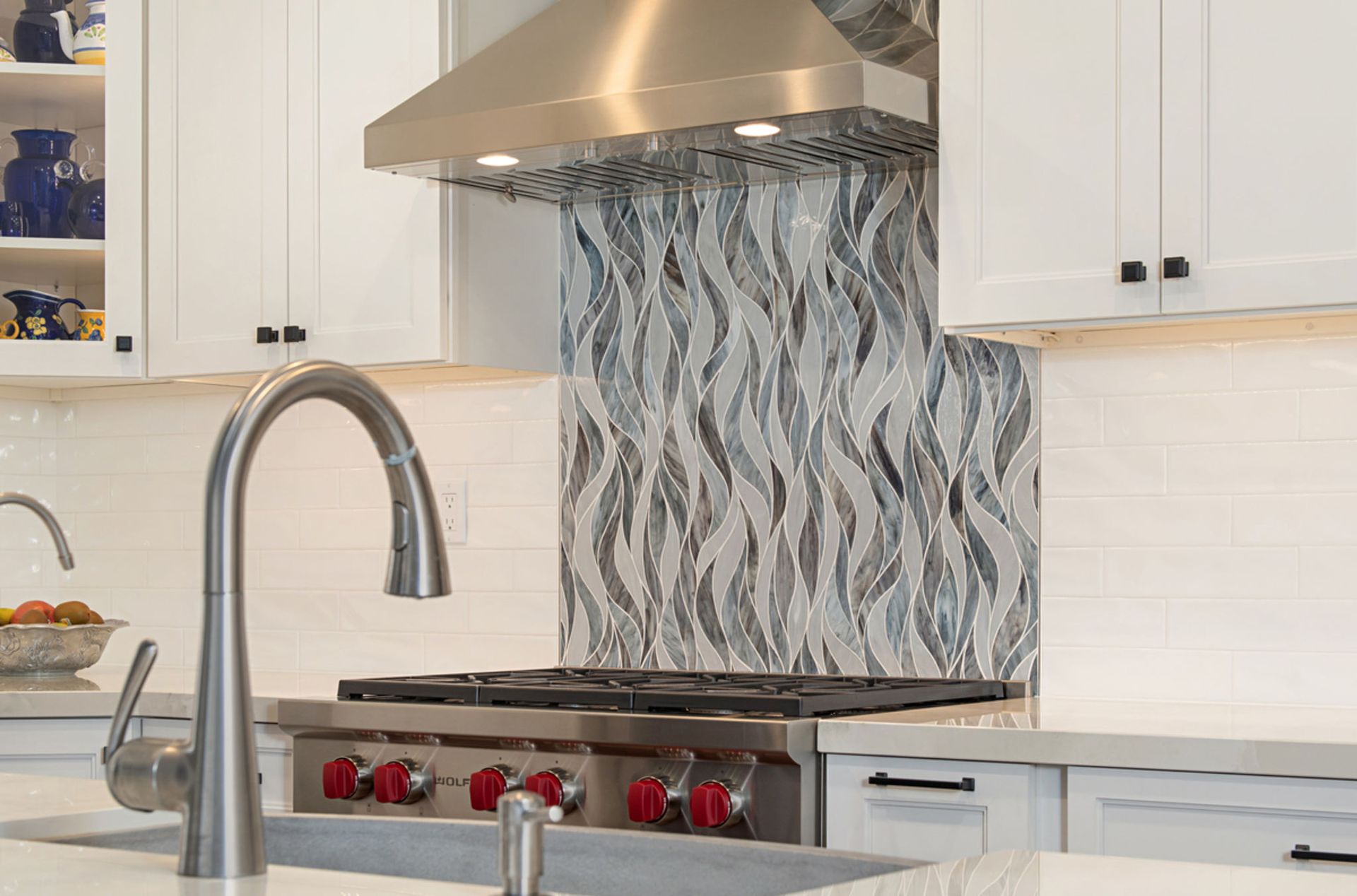 A modern kitchen with white cabinets, a stainless steel range, and a patterned backsplash. A faucet is in the foreground.