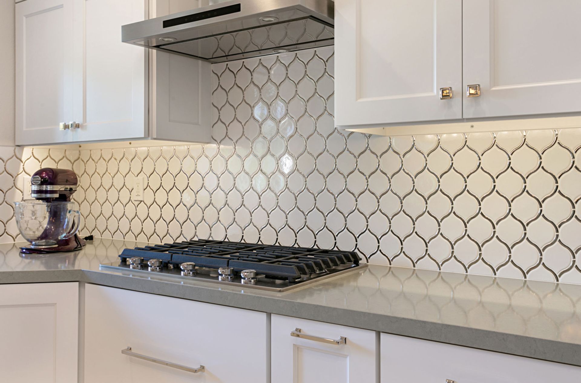 Kitchen with white cabinets, gray countertop, and patterned white tile backsplash. A gas stovetop is centered.