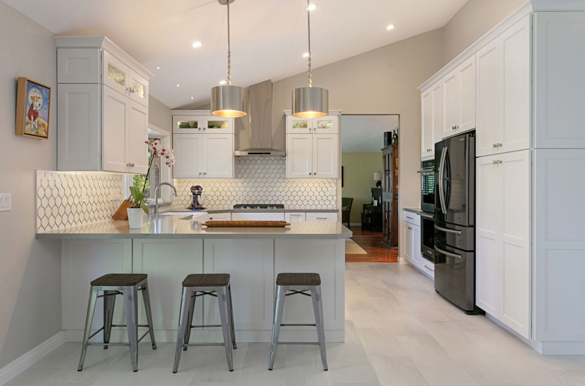 Modern kitchen with white cabinets, stainless steel appliances, and a light-colored countertop. Three barstools sit at the counter.