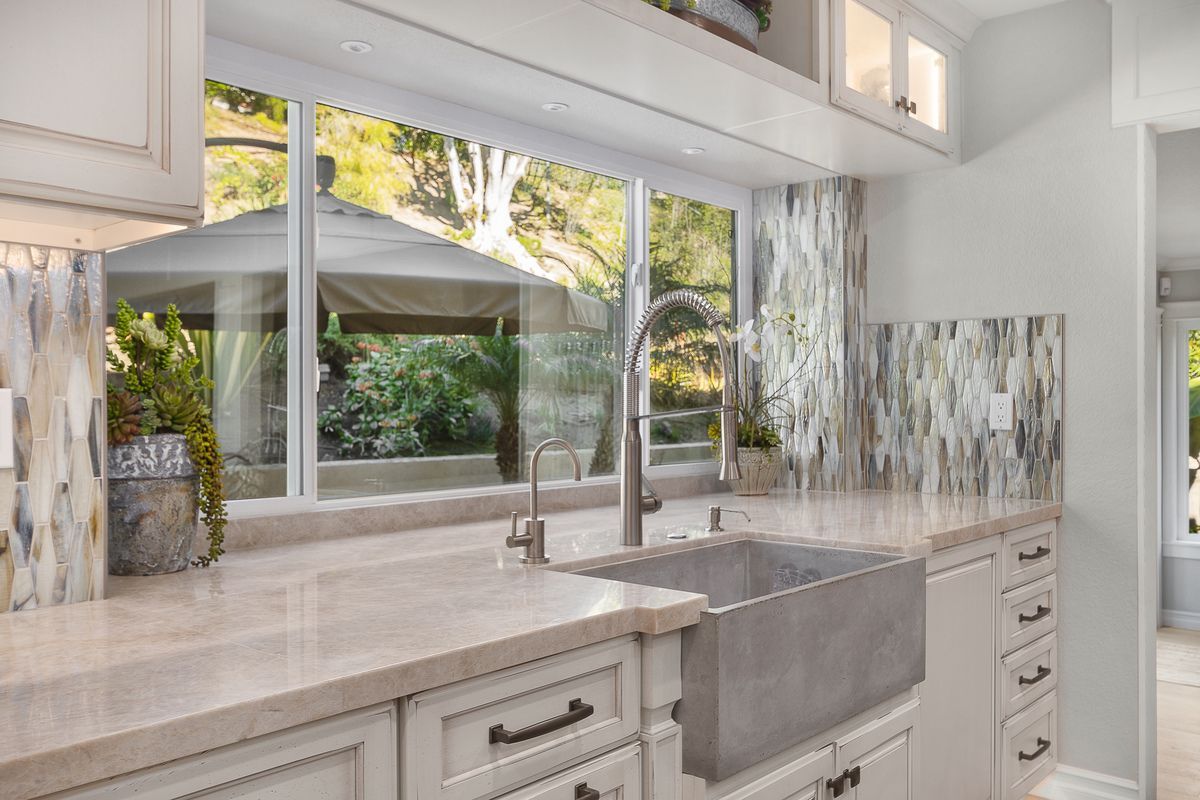 A bright kitchen with a stainless steel sink, light countertops, and a large window overlooking a garden.