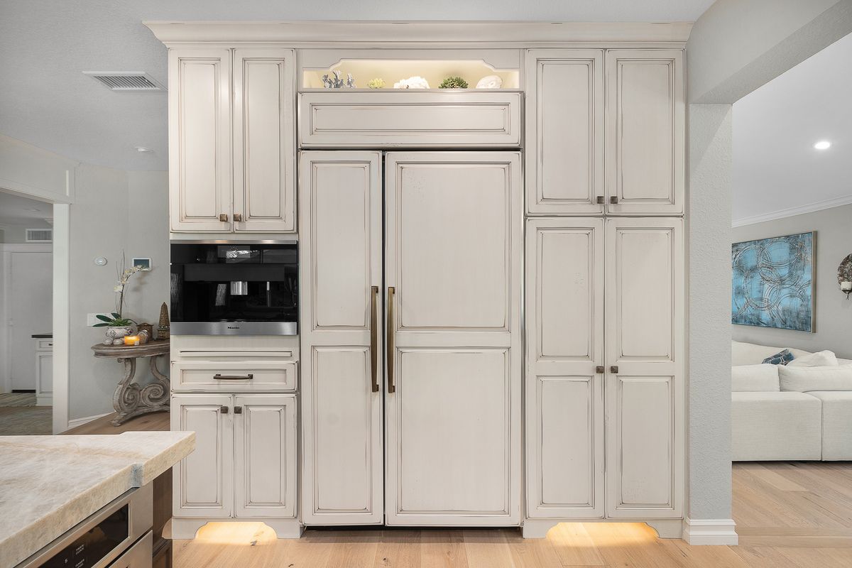 White kitchen cabinets with a built-in refrigerator and coffee maker. The cabinets are trimmed with light brown, and the kitchen is open to a living area.