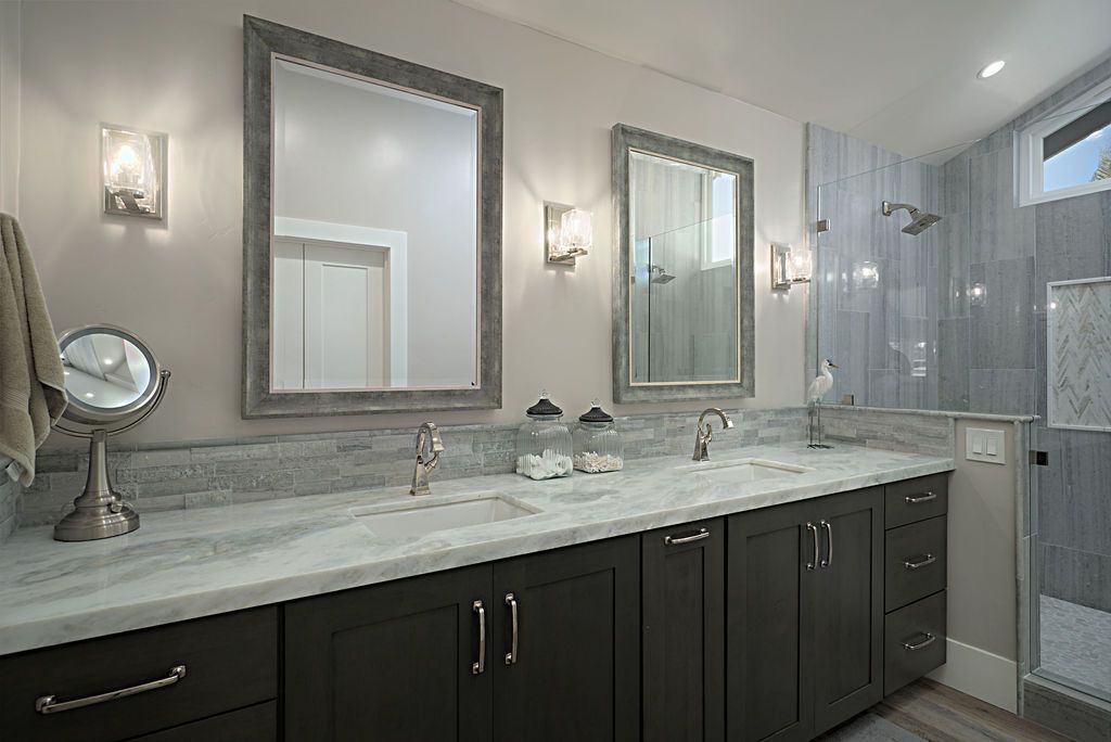 A modern bathroom with a double vanity, dark cabinets, marble countertop, two large mirrors, and a tiled shower.