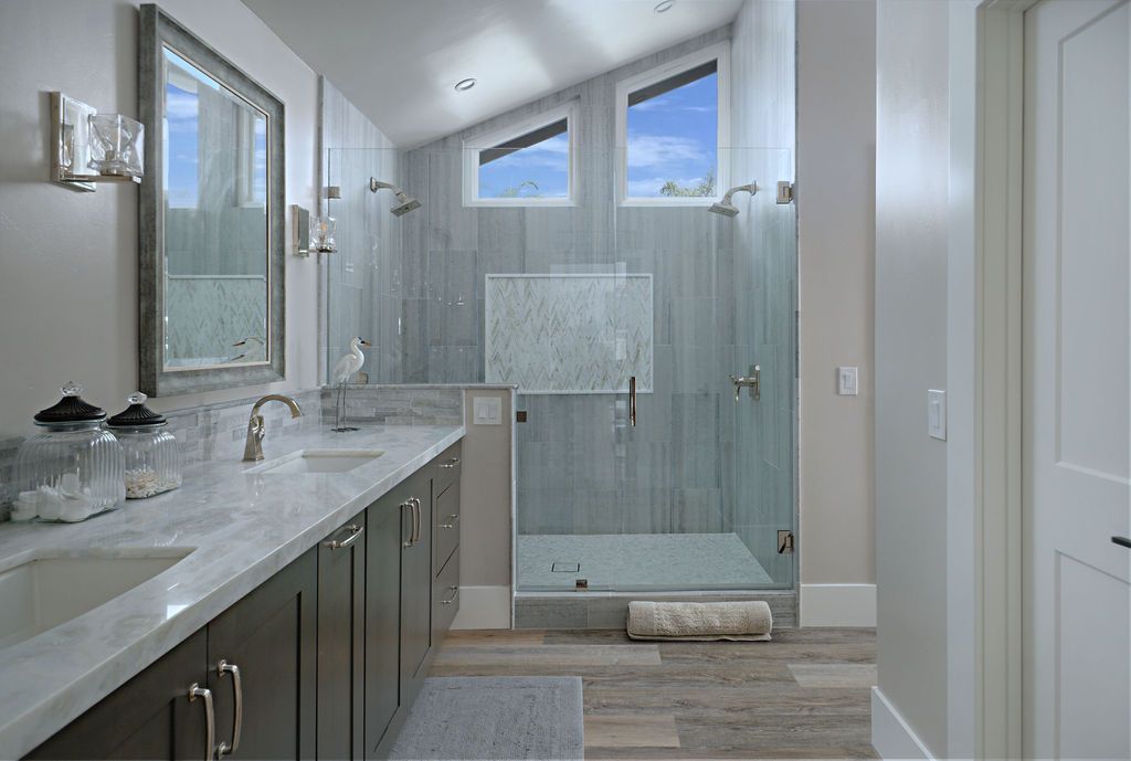 Bathroom with a double vanity, large glass shower, and three angled windows. Gray and wood tones dominate the space.