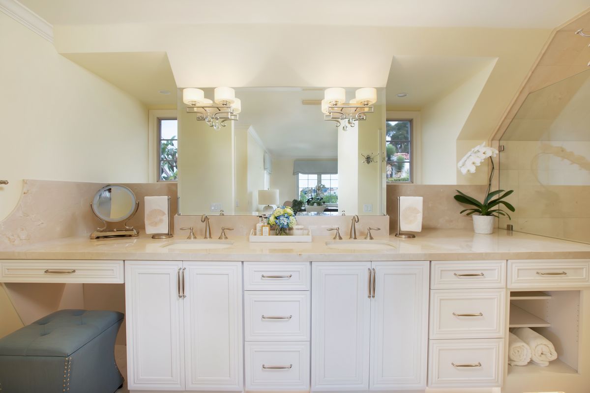 White bathroom vanity with double sinks, large mirror, and crystal chandeliers. Features white cabinets, beige countertops, and a blue ottoman.