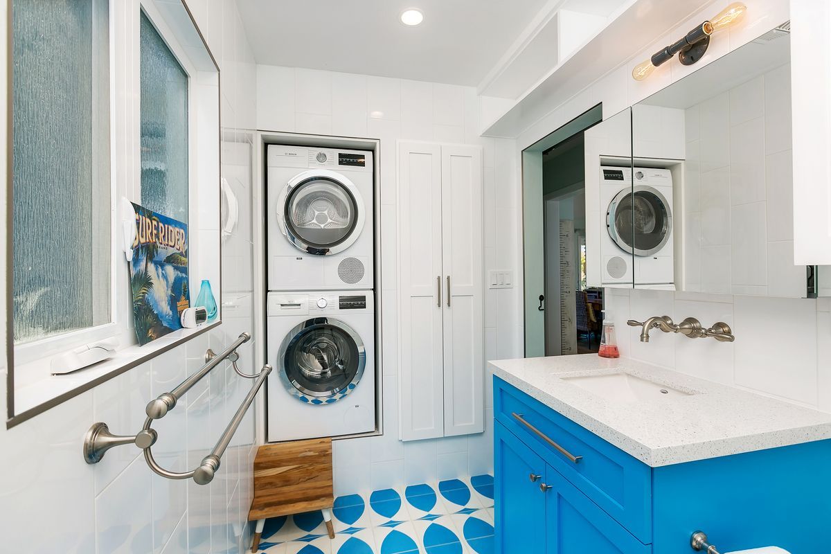A modern laundry room with a stacked washer and dryer, blue vanity, and patterned floor tiles. A window is on the left and a doorway is in the center.