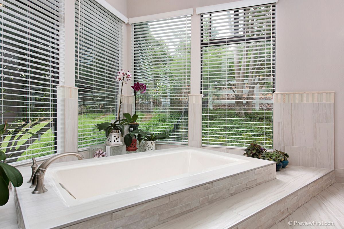 A bathroom with a built-in bathtub, surrounded by large windows with blinds, and plants.
