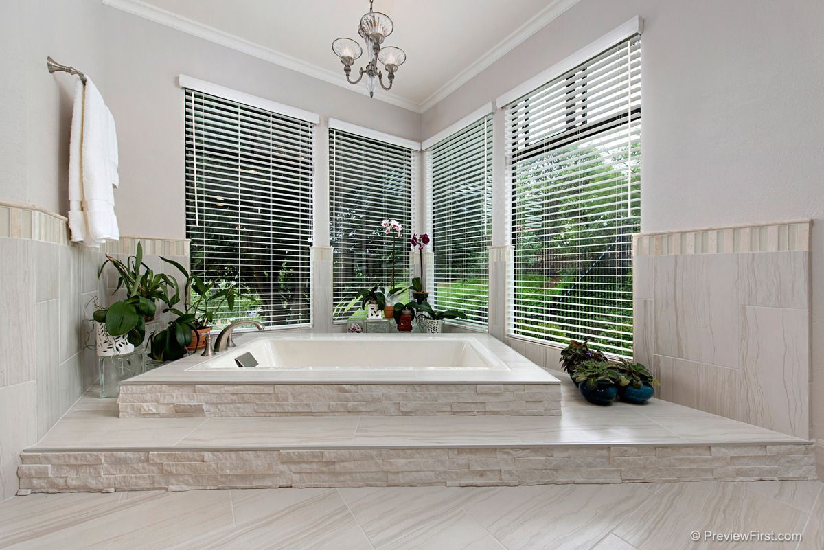 A light-filled bathroom with a raised bathtub in front of windows with blinds. Plants and a chandelier are visible.