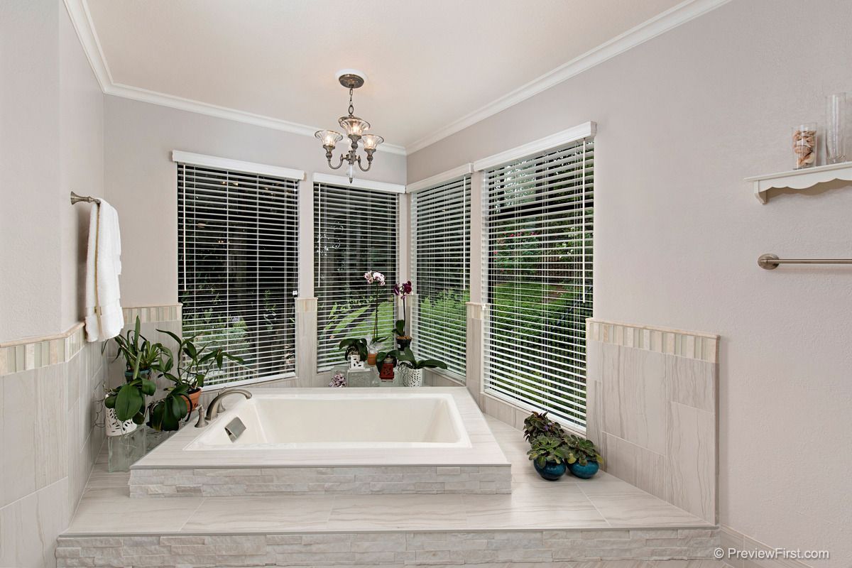 Bathroom with a white bathtub, surrounded by light-colored tile. Large windows with blinds and a chandelier illuminate the space.
