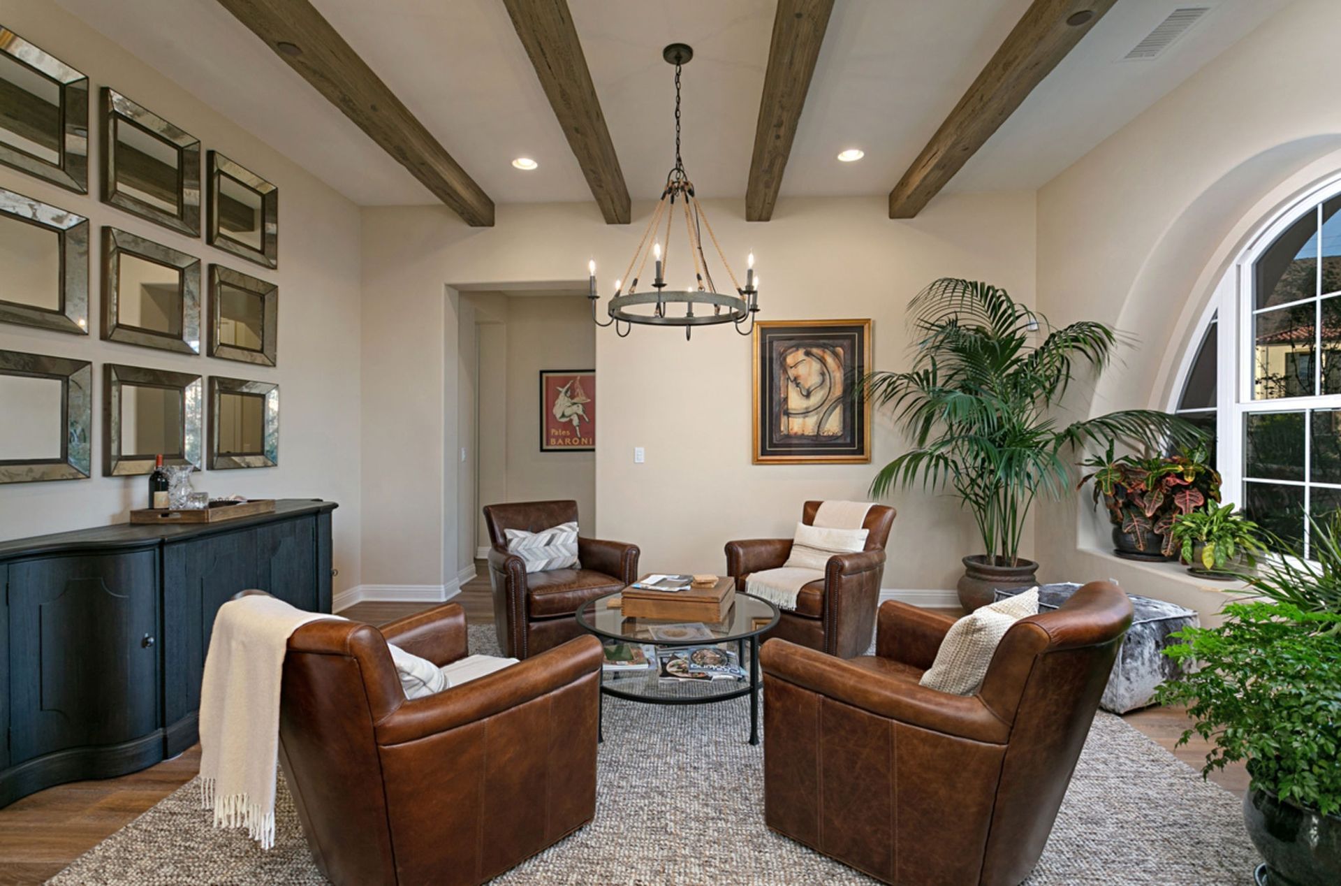 Cozy living room with four brown leather armchairs around a glass-topped table, under a chandelier. Features include wooden beams, mirrors, and a large arched window.
