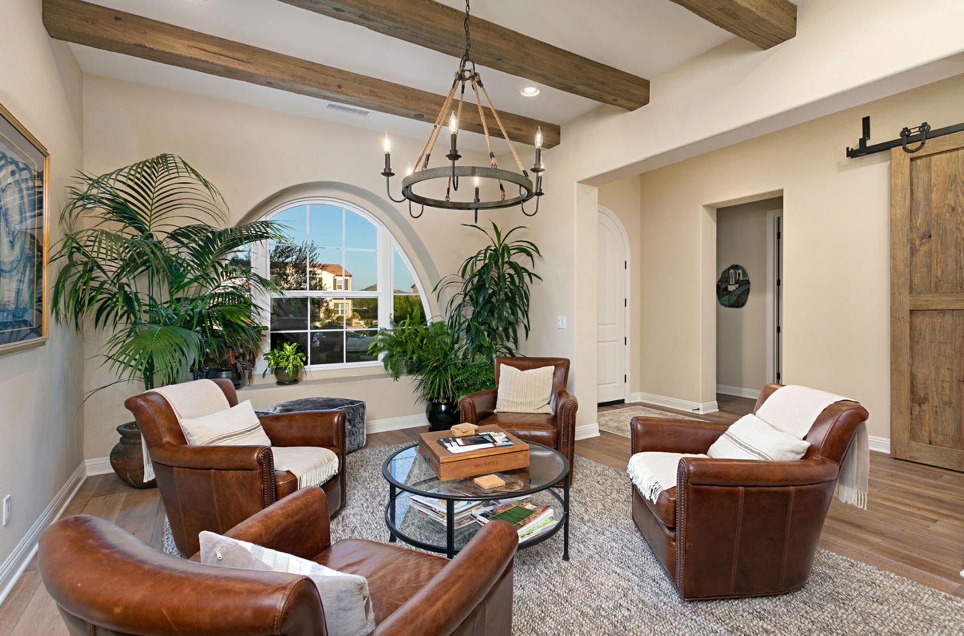 Living room with brown leather chairs arranged around a glass-topped coffee table, with arched window and potted plants.