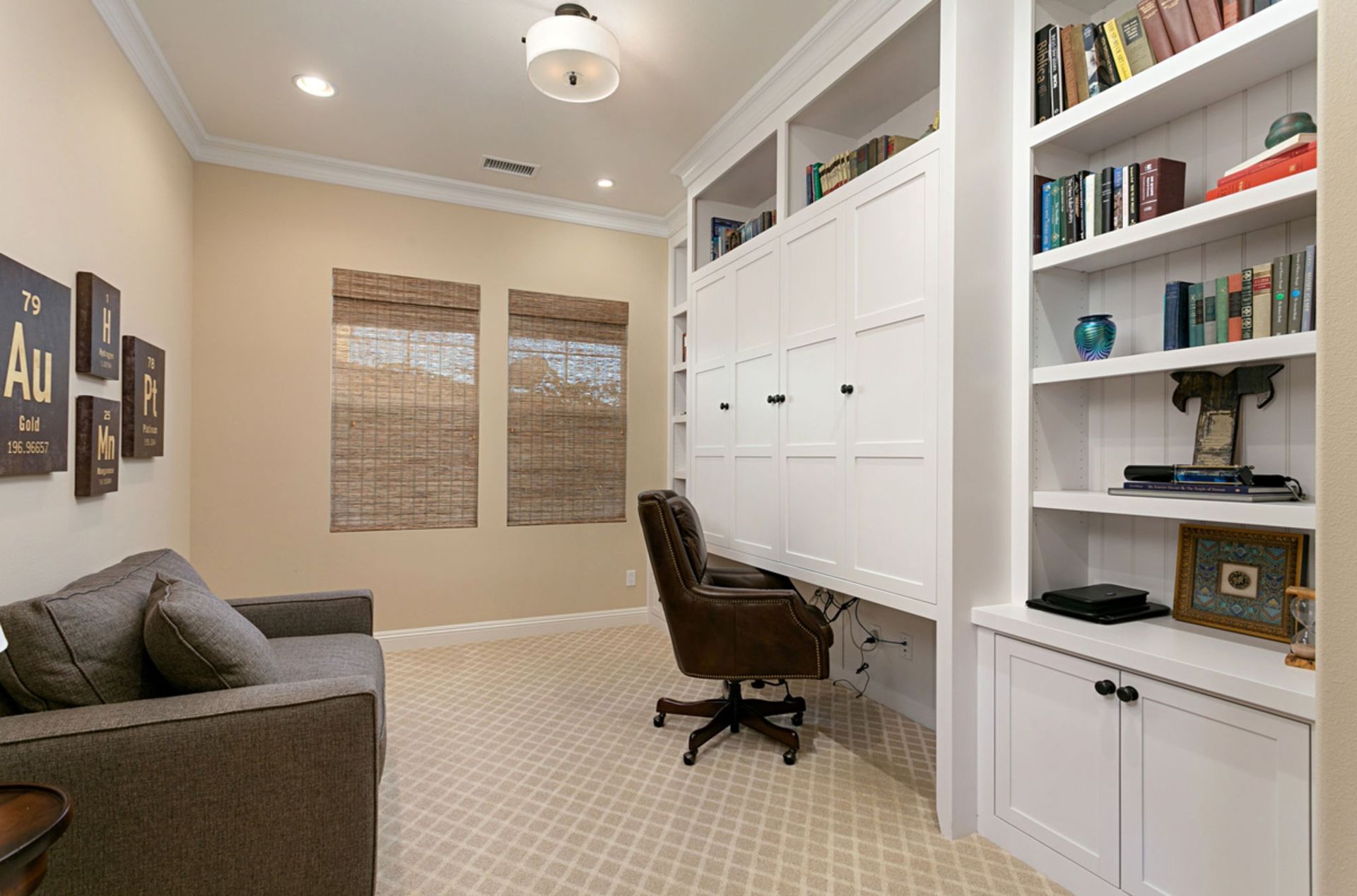 A home office with a built-in desk, bookshelves, sofa, and dark brown office chair; neutral tones throughout.