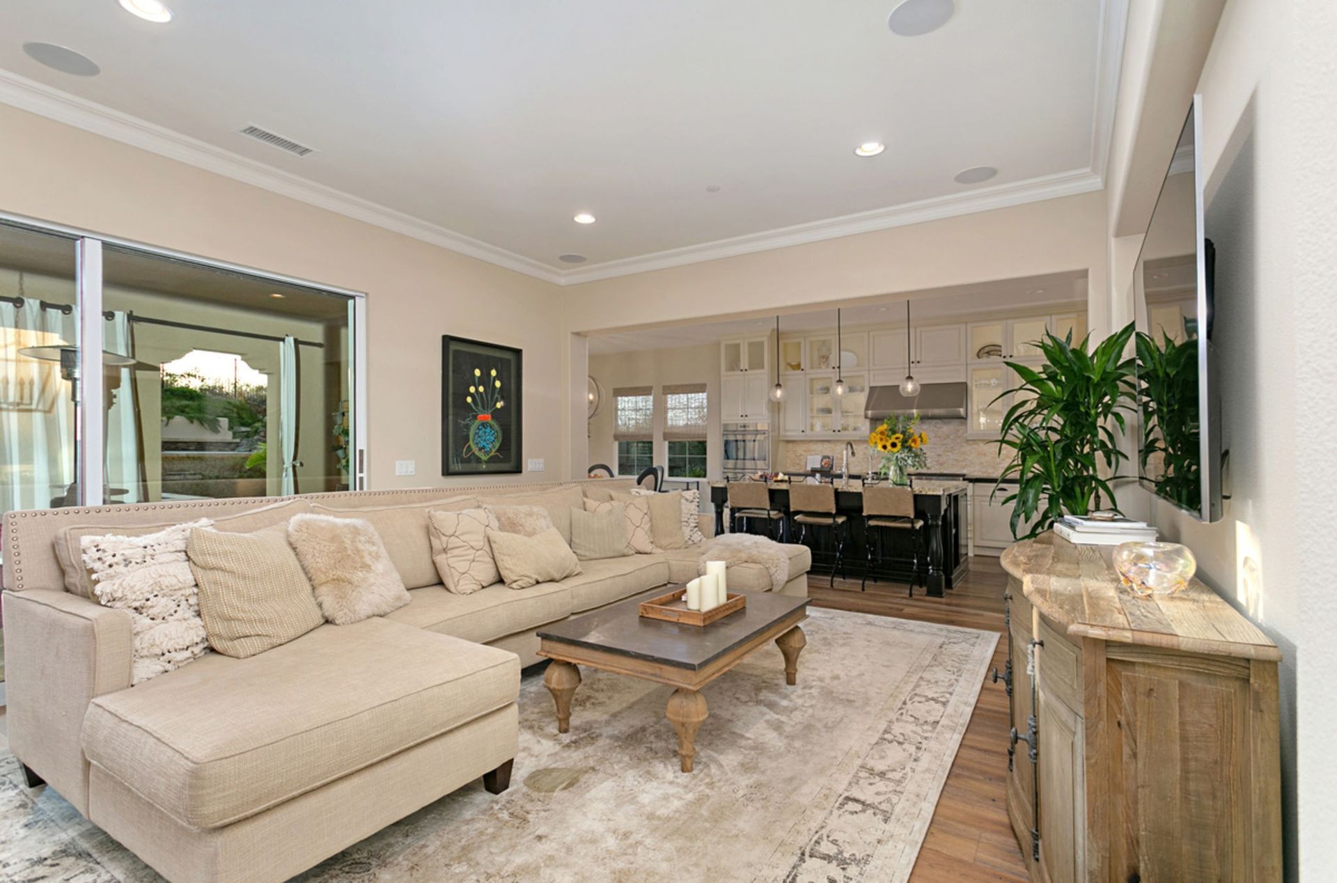 A bright living room with a sectional sofa, coffee table, and TV, leading to a kitchen with a dark island.