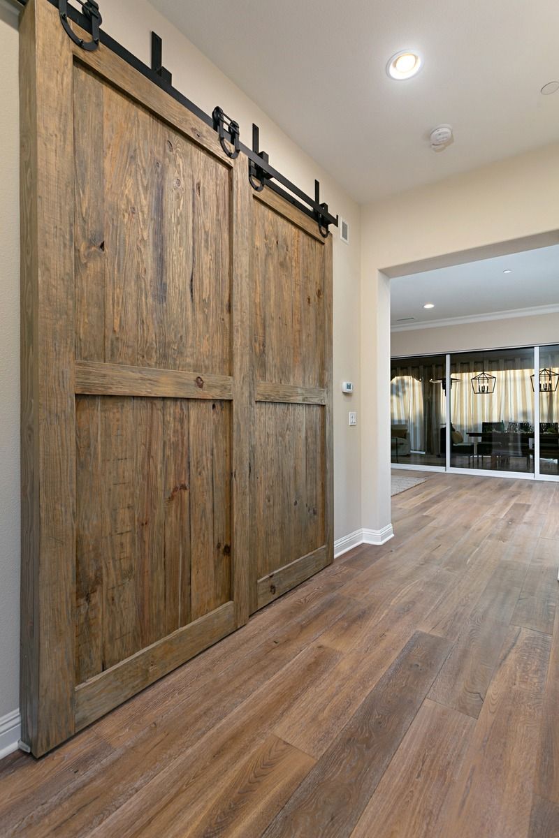 Two rustic wooden barn doors on a track system, set in a hallway with wood-look flooring leading to a room with glass walls.