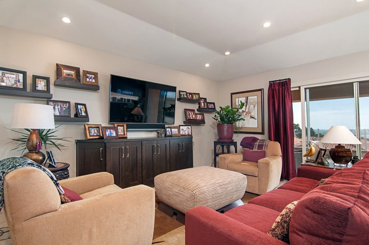 Living room with a red couch, two tan armchairs, and a patterned ottoman.  A large TV is mounted above a dark wood cabinet; framed photos line the walls.