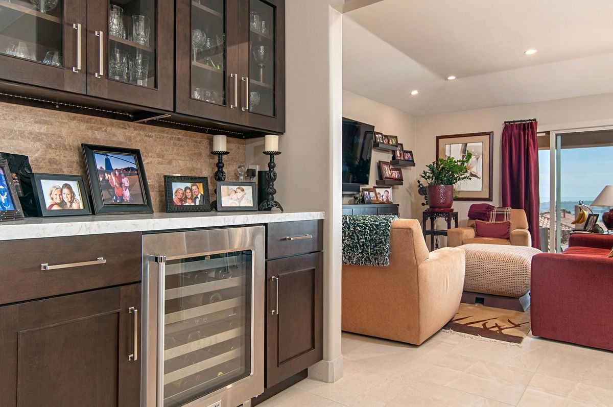 A built-in bar with a wine fridge, dark cabinets, and framed photos leads into a living room with a view of the ocean.