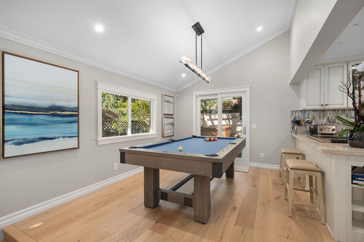 Pool table in a light-filled room with hardwood floors, a painting, and a bar area. Daylight streams in from a window and sliding glass door.
