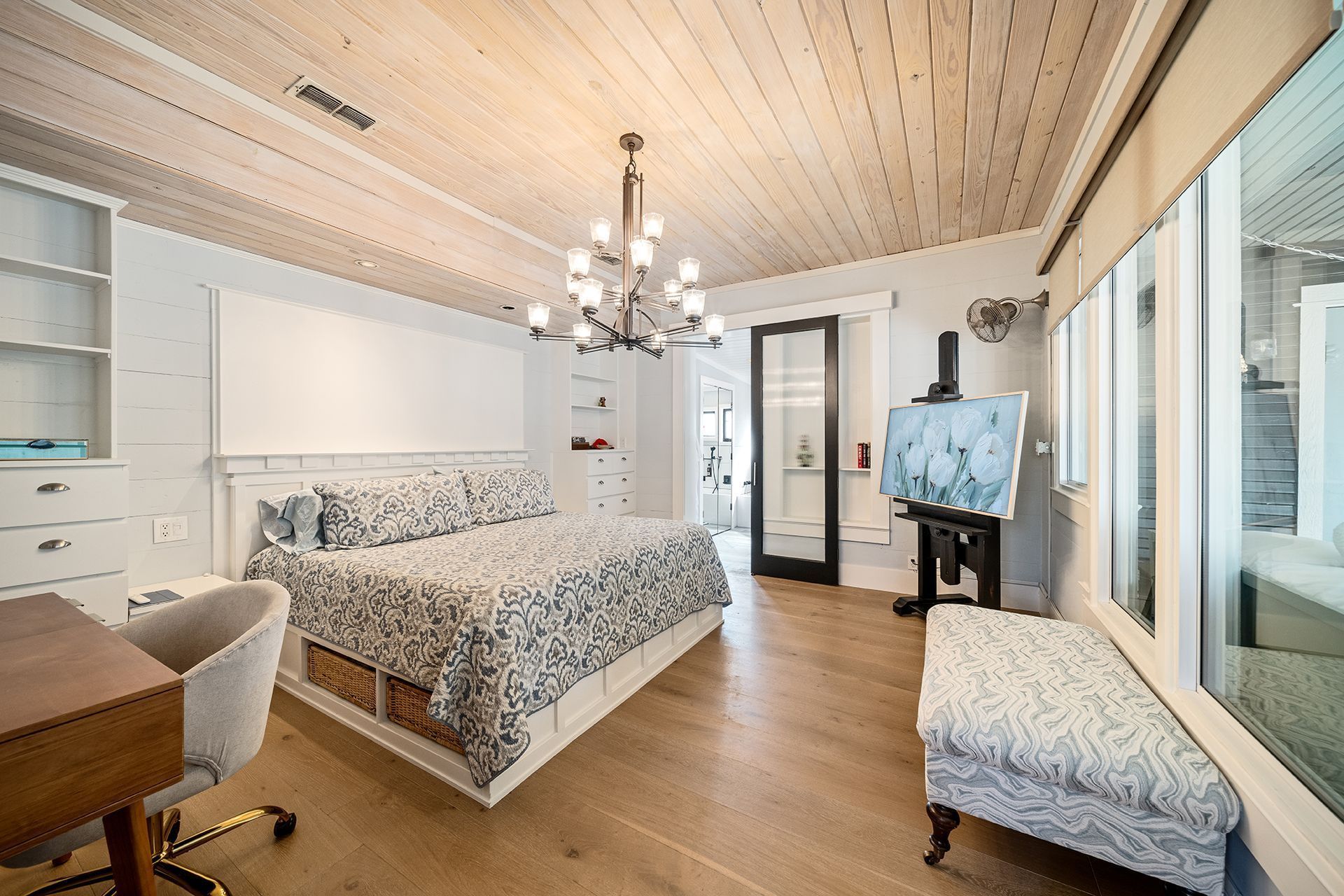A bright bedroom with a bed, desk, and artwork. The room has a light wood ceiling and floor, with white walls.