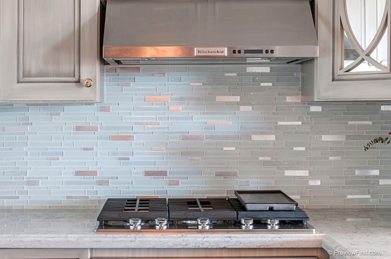 A modern kitchen backsplash with a gas stove, stainless steel range hood, and light blue and silver tile.