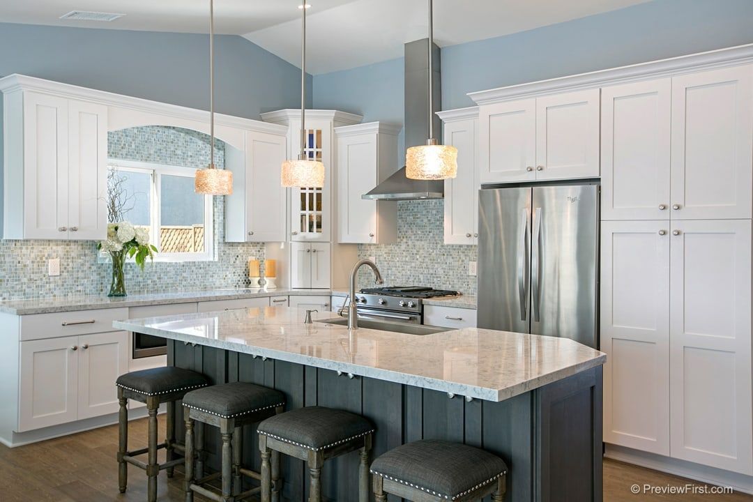 Modern kitchen with white cabinets, blue walls, and a gray island with stools. Stainless steel appliances and pendant lights.