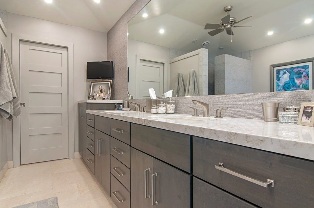 Modern bathroom with a large mirror, gray cabinets, double sinks, and a small TV above the countertop.