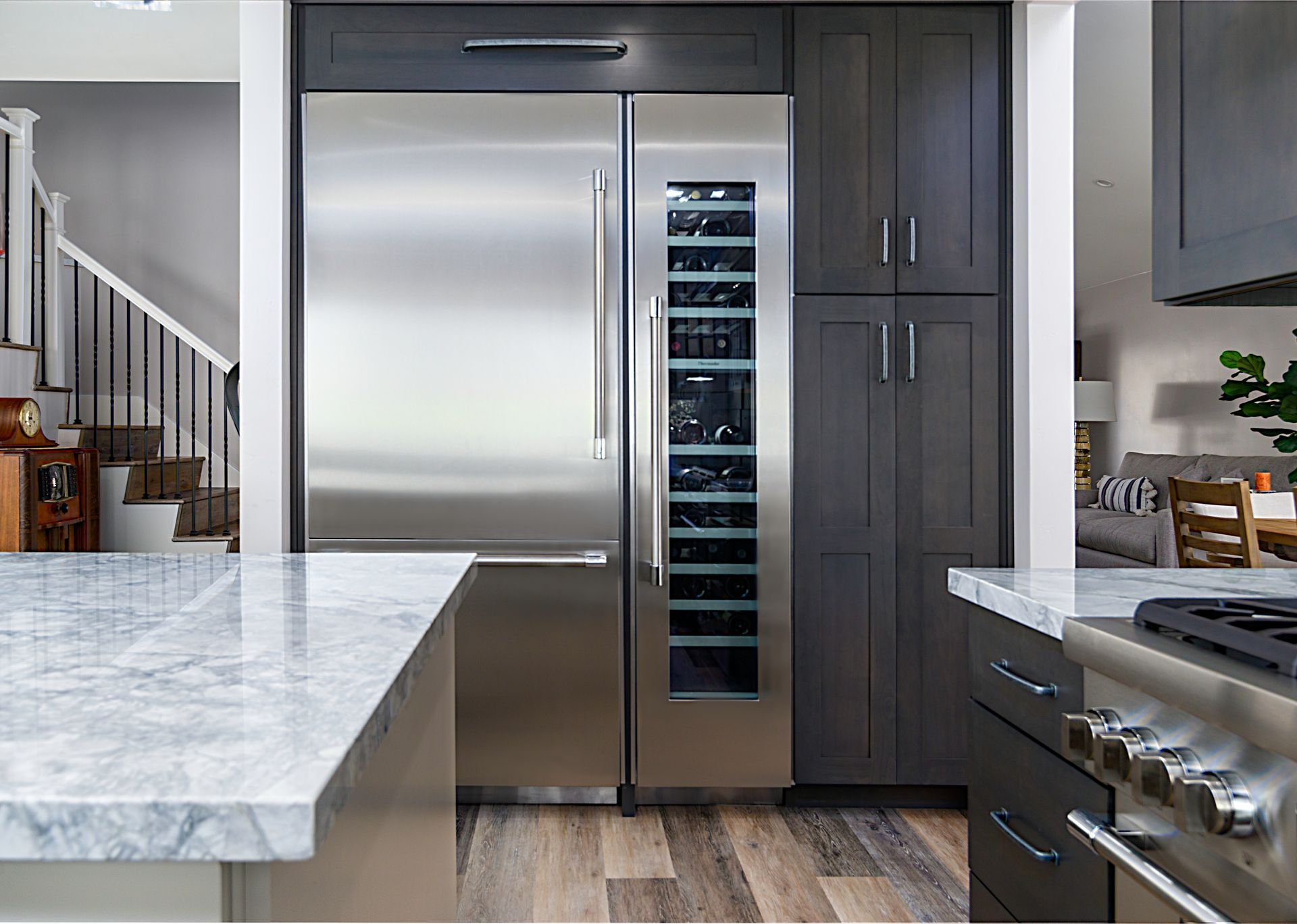 Modern kitchen featuring a stainless steel refrigerator and wine cooler framed by dark gray cabinets. Marble countertops and wood flooring complete the space.