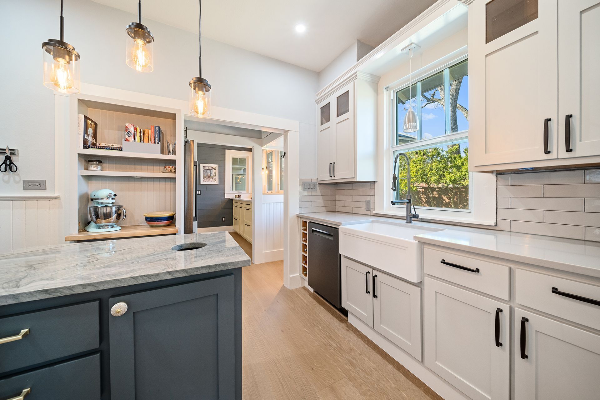 Kitchen with white and navy blue cabinetry, a farmhouse sink, and a granite countertop island, light wood flooring, and hanging light fixtures.