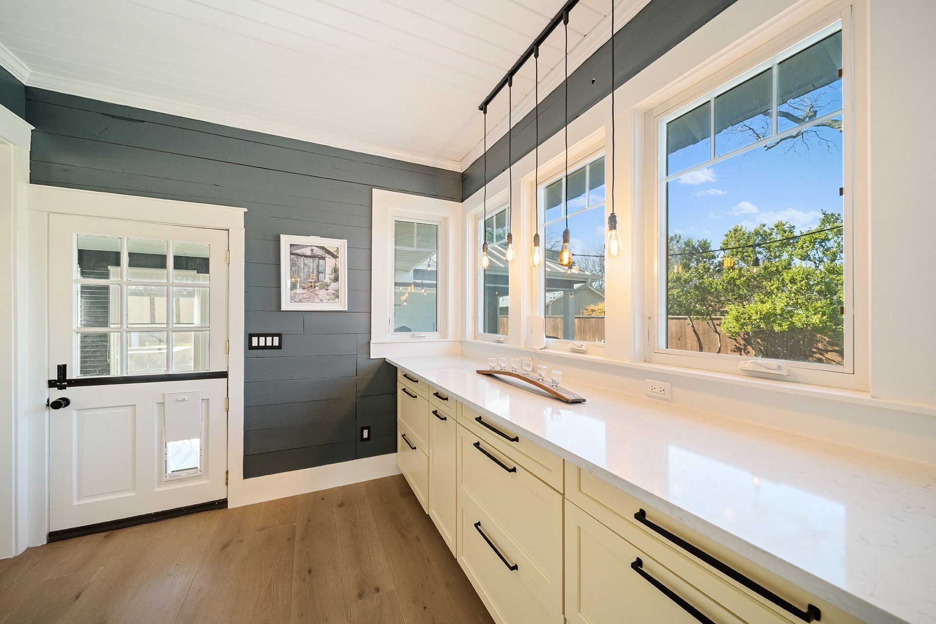 Kitchen with white cabinets, countertops, and trim, dark gray wall, and three windows overlooking a bright, sunny outdoor scene.