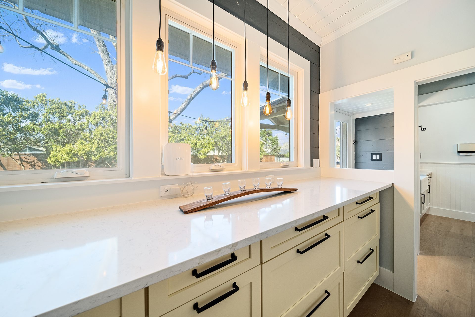 Cream-colored cabinets line a bright room with a white countertop and three pendant lights. Windows offer a view of trees and a clear sky.