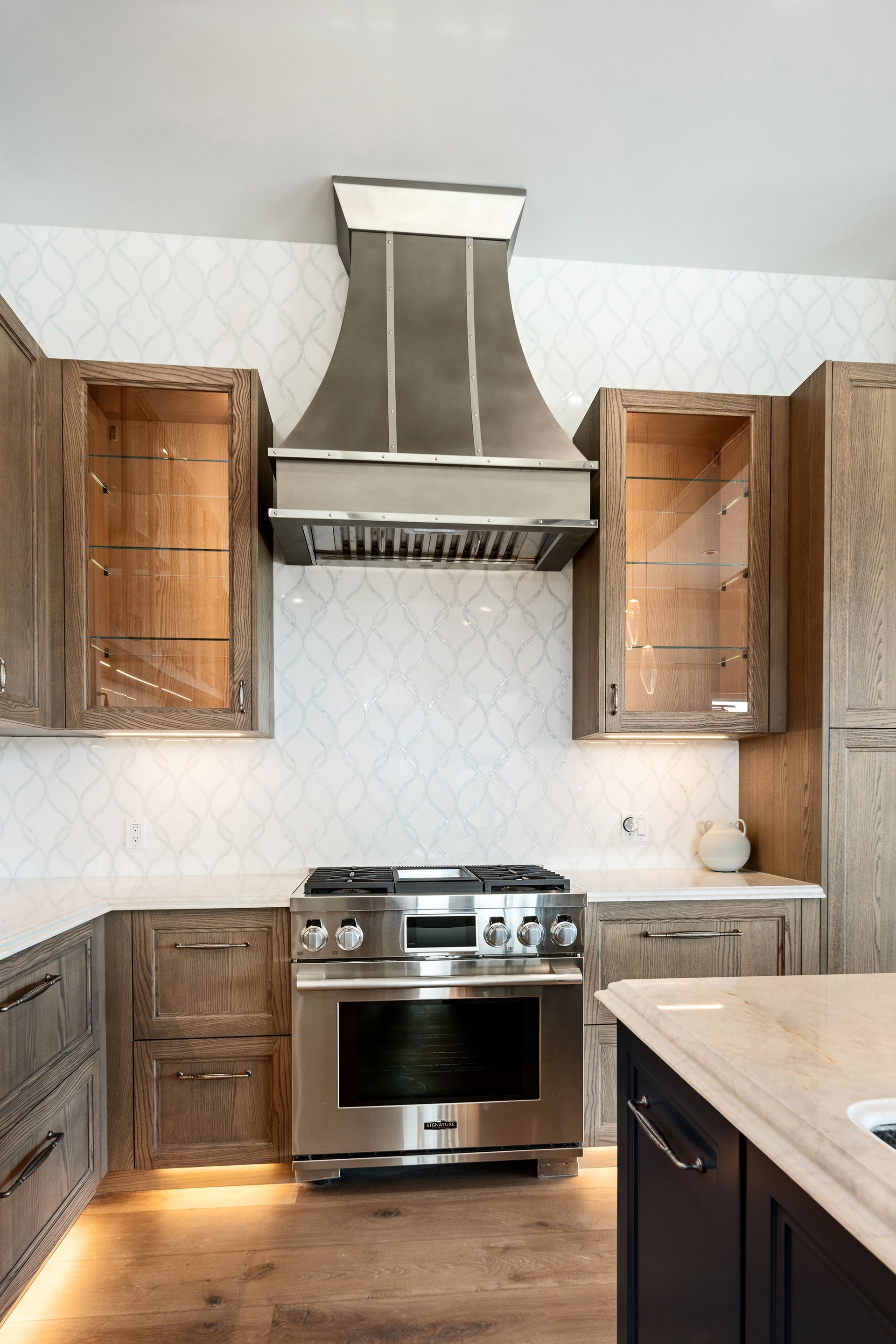 Modern kitchen with stainless steel appliances, wooden cabinets, and a light-colored countertop. The range hood is prominent against a patterned backsplash.
