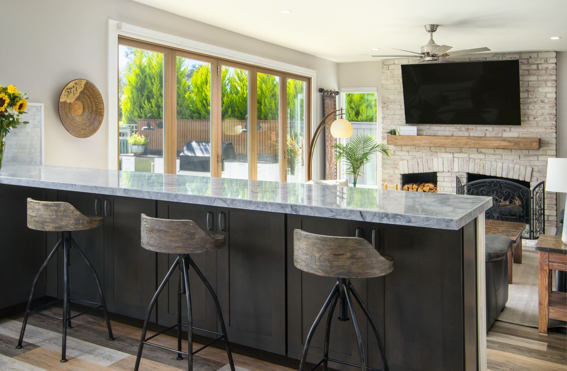 Modern kitchen with a bar, featuring stools, and a view of a patio through large windows. The living room includes a fireplace and TV.