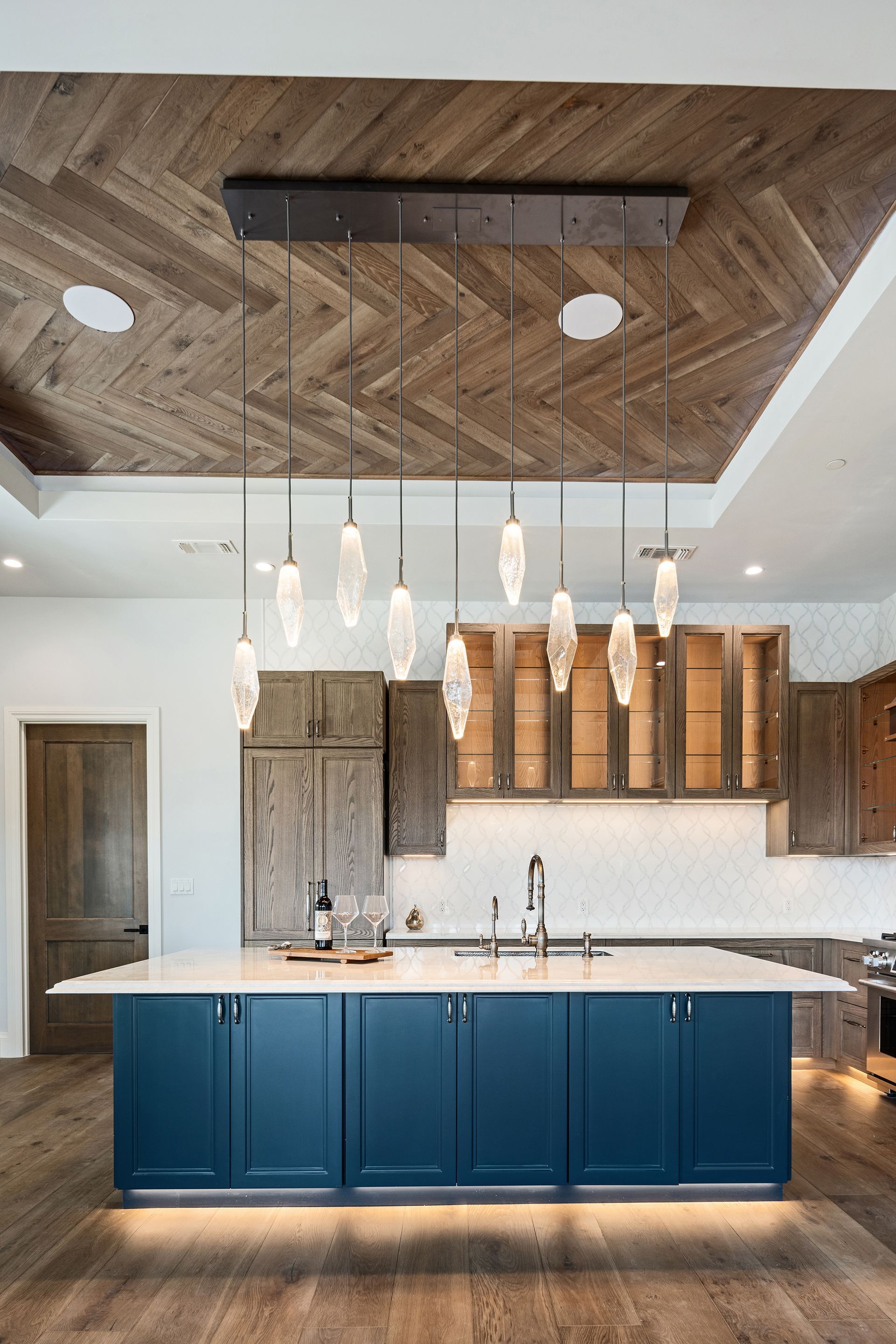 A modern kitchen with a blue island, wooden cabinets, and hanging crystal pendant lights; wooden herringbone ceiling.