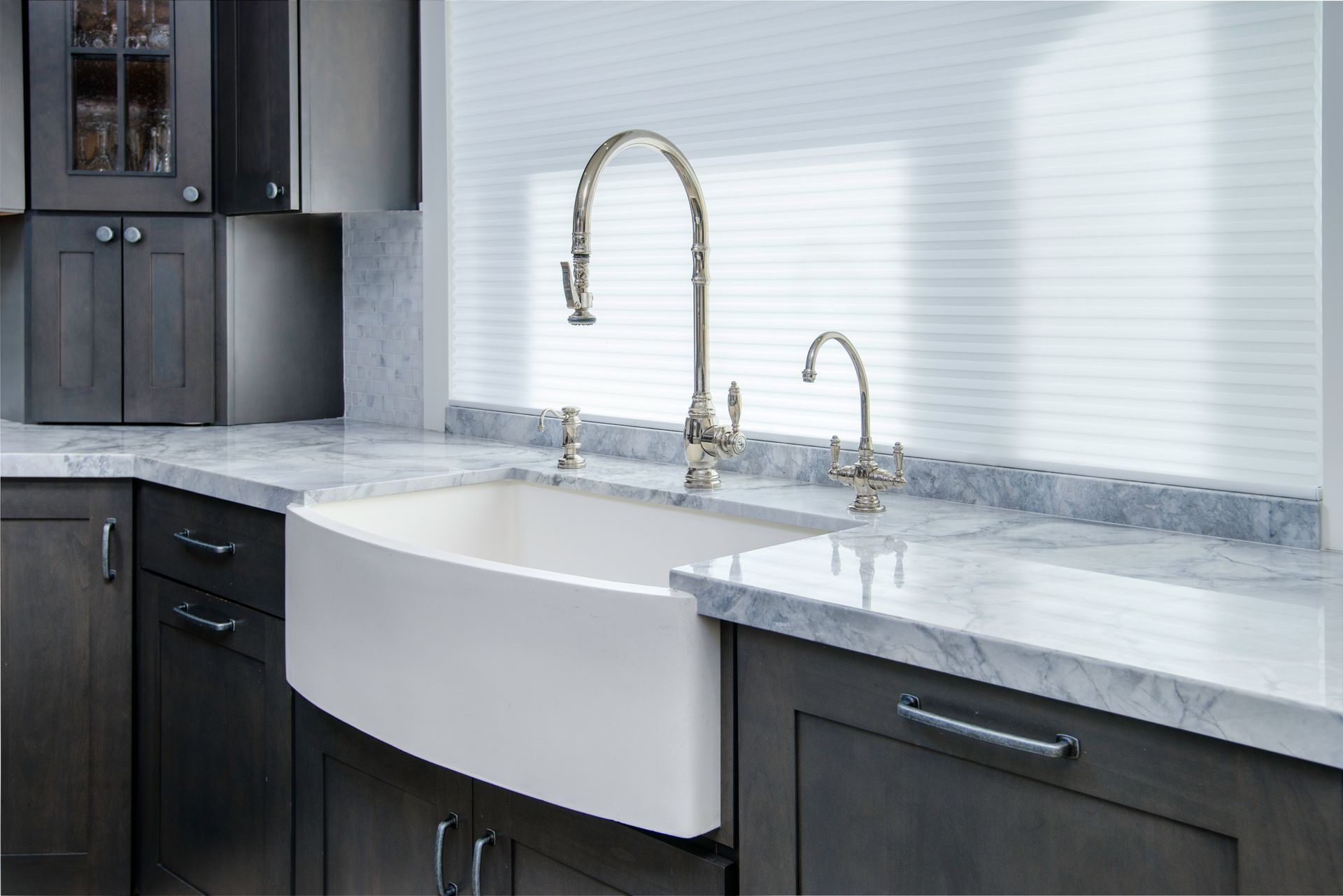 A farmhouse kitchen sink with a tall, curved faucet, set against a white countertop and dark gray cabinets.