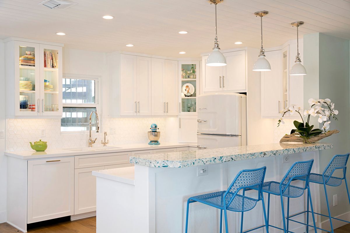 A bright white kitchen with blue accents, featuring white cabinets, blue bar stools, and pendant lights over a breakfast bar.