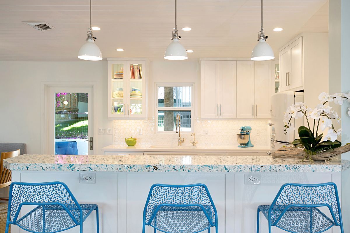 A bright, modern kitchen with white cabinets, a colorful speckled countertop, and blue bar stools. Three pendant lights hang above the countertop.
