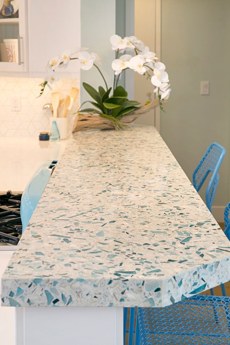 A kitchen counter with white and teal speckled surface, adorned with a white orchid arrangement. Two blue chairs are placed at the counter.