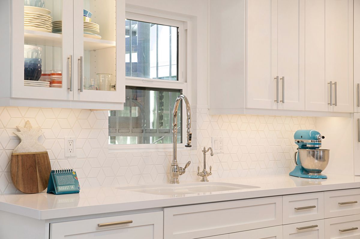 Bright white kitchen with cabinetry, a stainless steel faucet, and a light blue mixer. The kitchen has white countertops, a window, and a white backsplash.