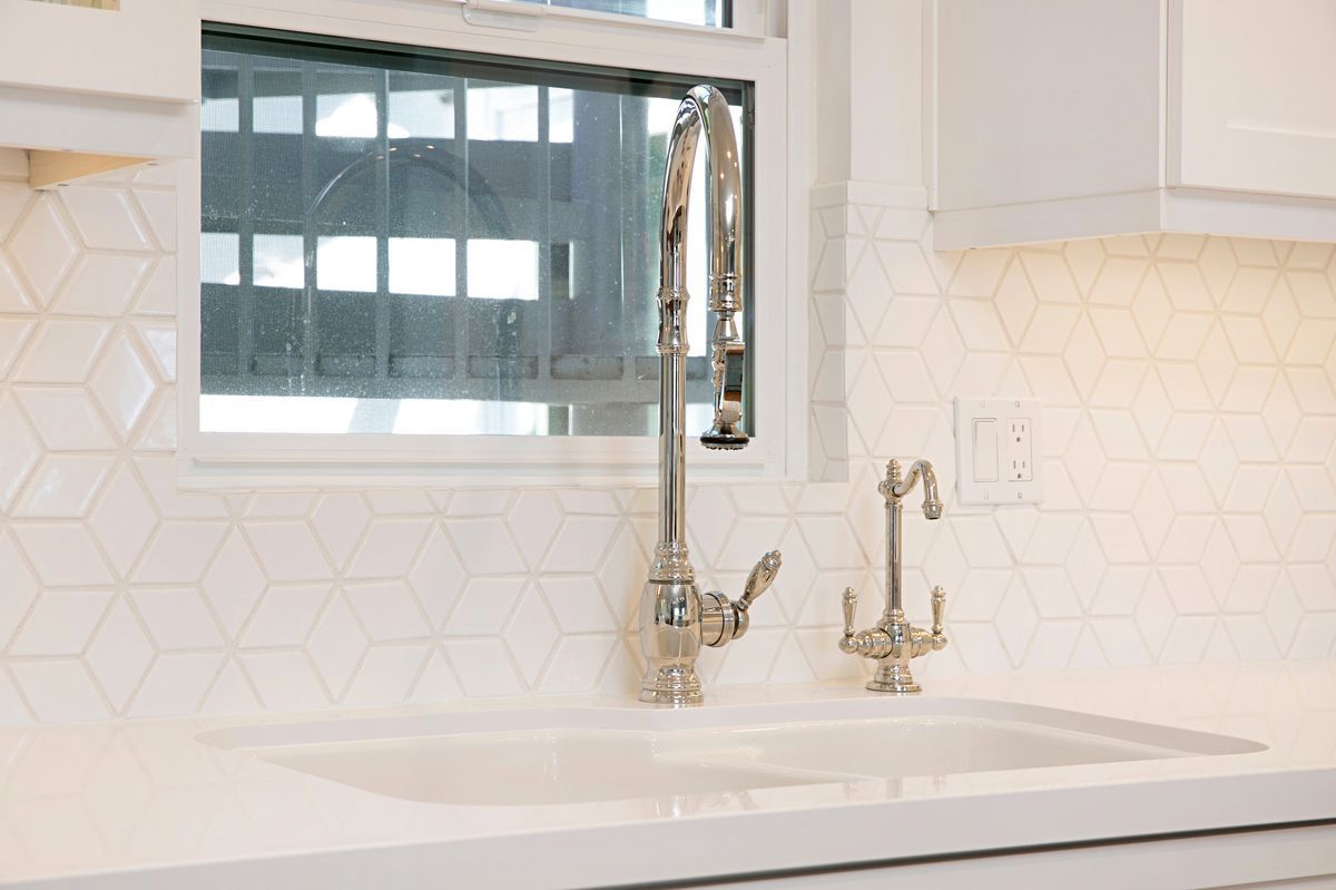 White kitchen sink with chrome faucet and geometric tile backsplash, in front of a window.