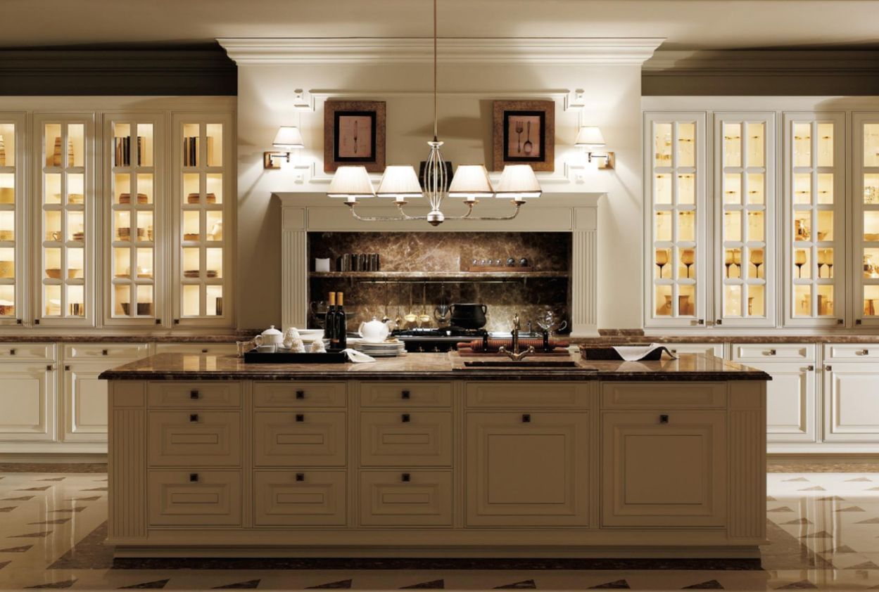 A luxurious kitchen with cream-colored cabinets and a large island. Glass-fronted cabinets are illuminated, and a chandelier hangs above.