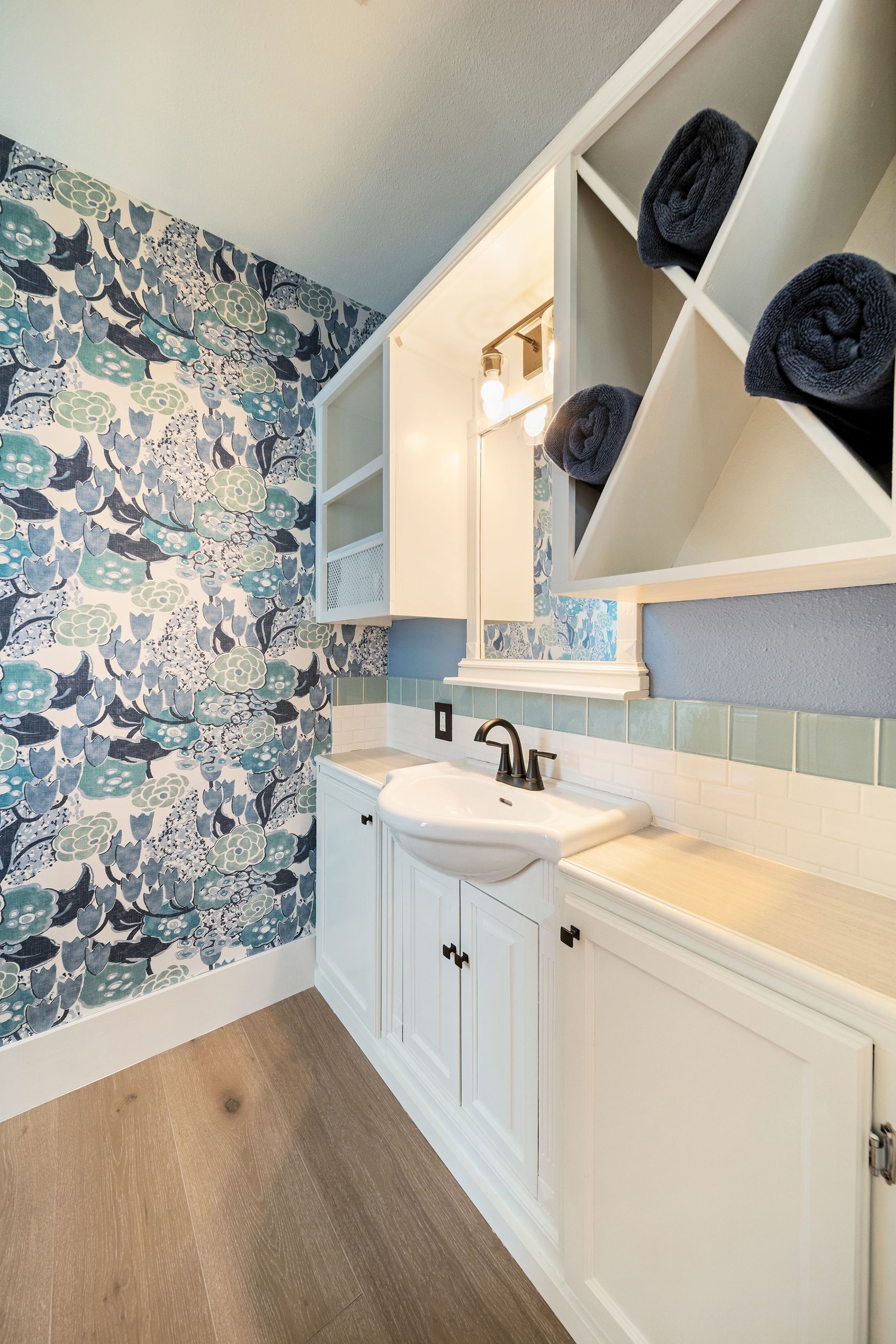 A small white bathroom with blue wallpaper, a sink, cabinets, and built-in shelving with rolled towels.