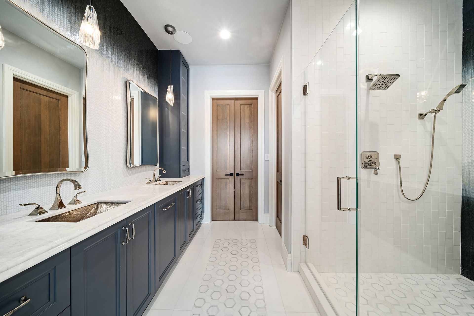 Modern bathroom with navy blue cabinets, white tile, and a glass shower. A long vanity and double doors at the end.