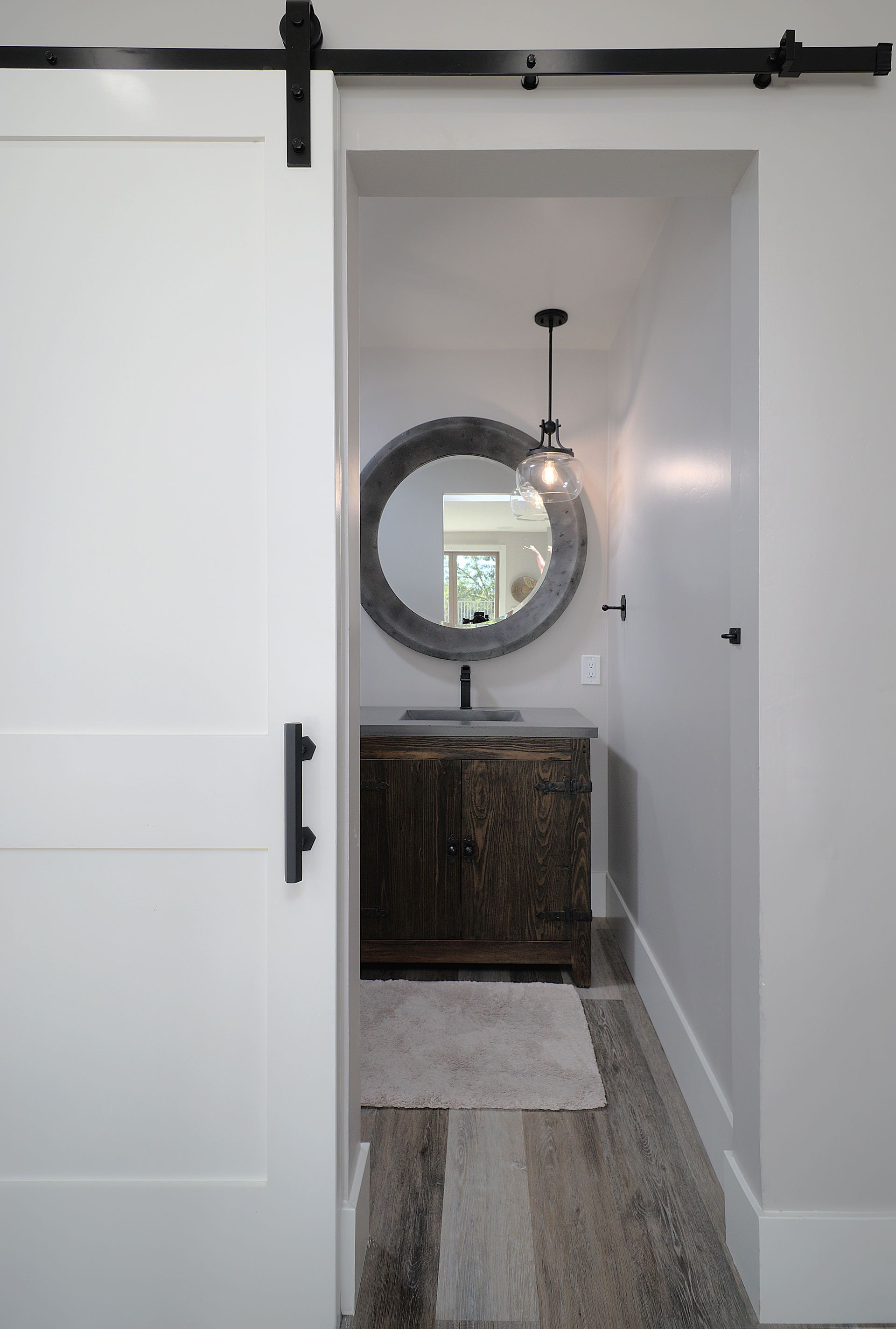 White barn door open to a narrow bathroom with a rustic vanity, round mirror, and pendant light.
